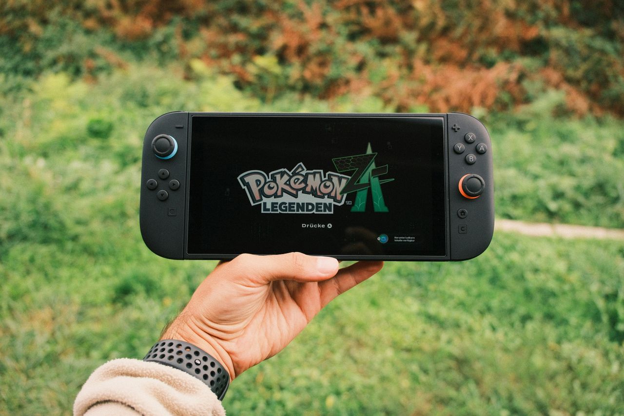about-us A hand holding a handheld gaming console displaying the Pokémon Legends game screen outdoors.